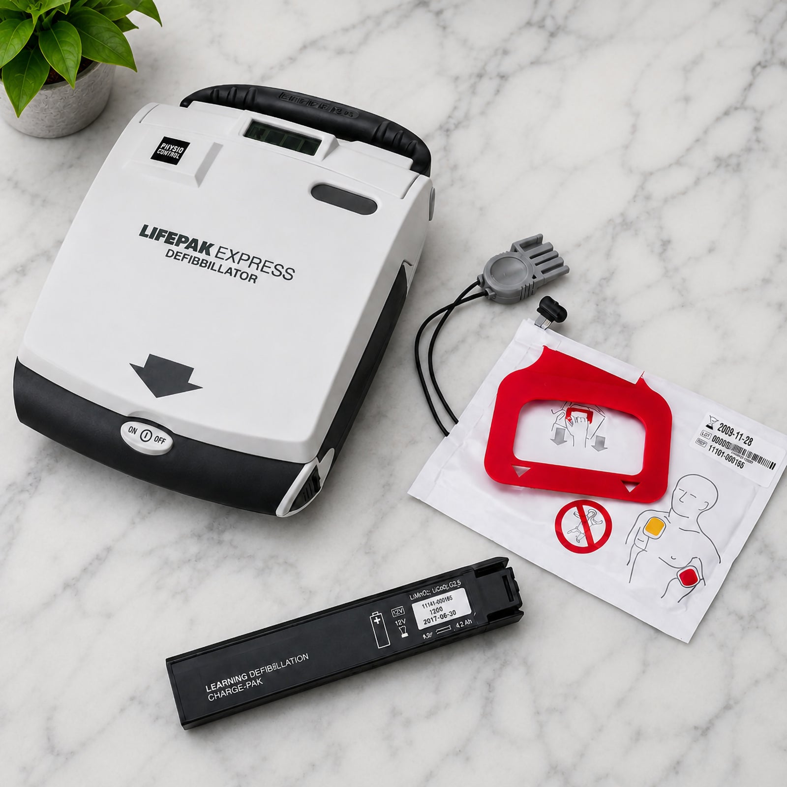 LifePak CR Plus Charge Pak components displayed with a LifePak Express defibrillator on a clean marble surface. The top view shows the adult electrode pad package, connected pad assembly and replacement battery pack positioned beside the compatible AED, providing a clear visual reference for routine LifePak Express maintenance and replacement preparation.