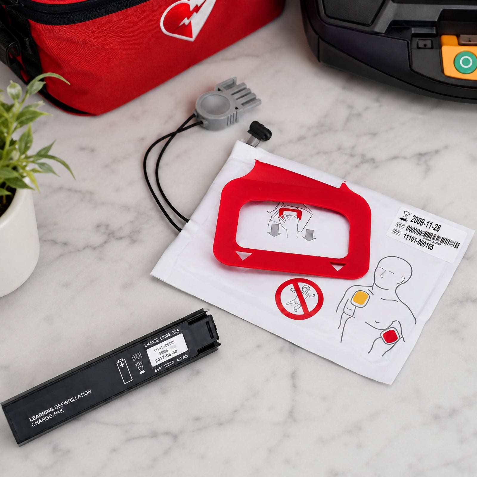 Close up lifestyle view of the LifePak CR Plus Charge Pak components on a marble surface, with the packaged adult electrode pads, connector and battery pack prominently displayed. A defibrillator and AED carry case appear partially in the background, creating a practical equipment maintenance setting while keeping attention on the replacement consumables.