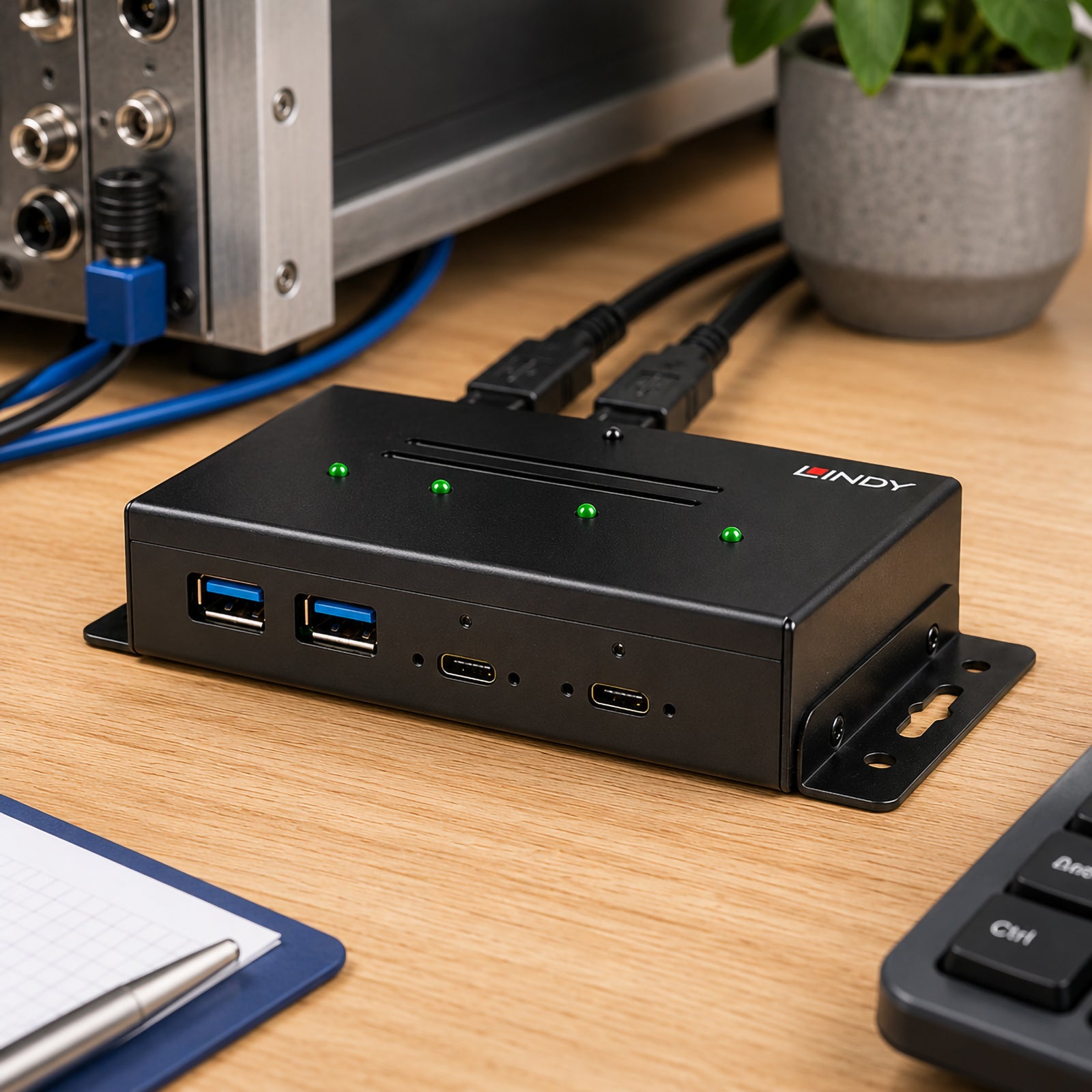 Front angled view of the Lindy industrial USB hub placed on a wooden workstation beside industrial equipment. The two USB Type A ports, two USB Type C ports, LED indicators and integrated mounting brackets are clearly visible, showcasing a professional solution for reliable USB expansion.