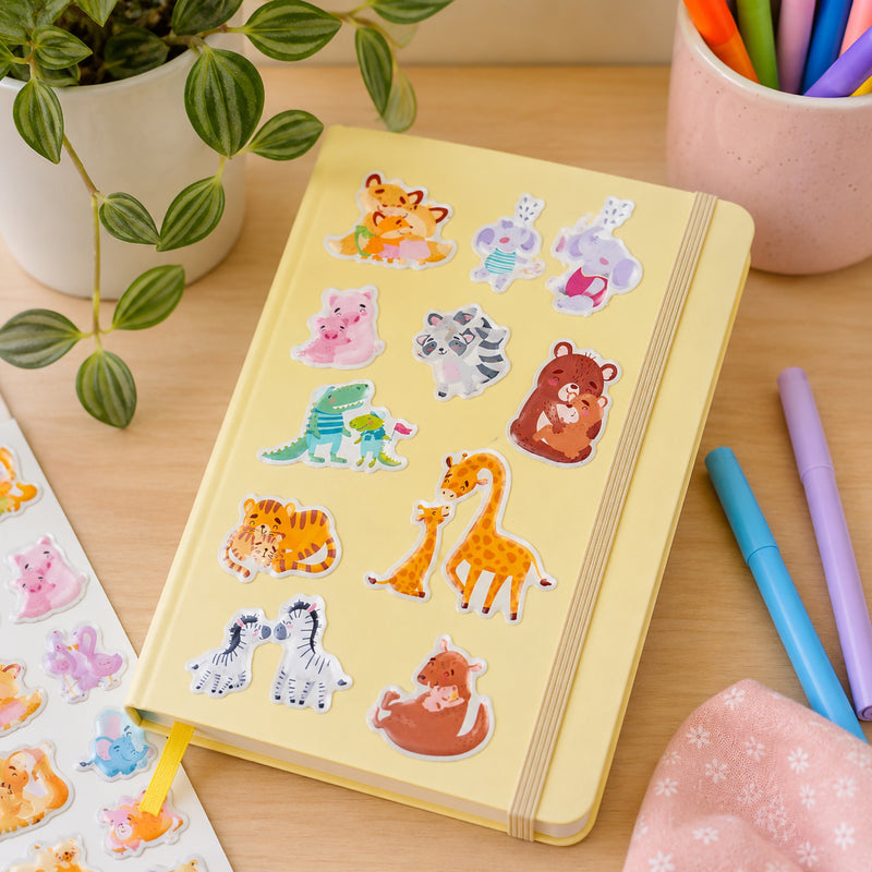 Yellow notebook personalised with adorable puffy animal stickers showing loving parent and baby animal pairs. Colourful pens and stationery surround the notebook, highlighting creative uses for decorating journals and personal items.