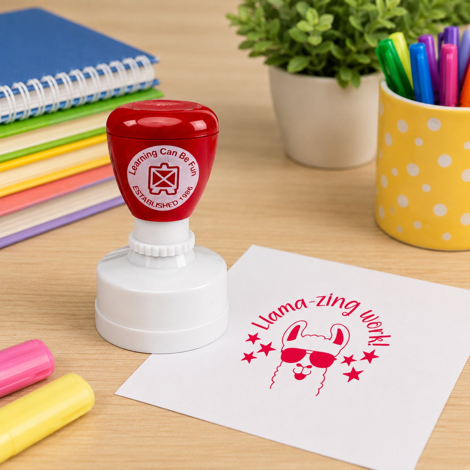 Learning Can Be Fun Llamazing Work Merit Stamp displayed on a classroom desk beside colourful markers and notebooks. The bright red llama themed impression showcases a fun motivational message that celebrates outstanding student achievement.