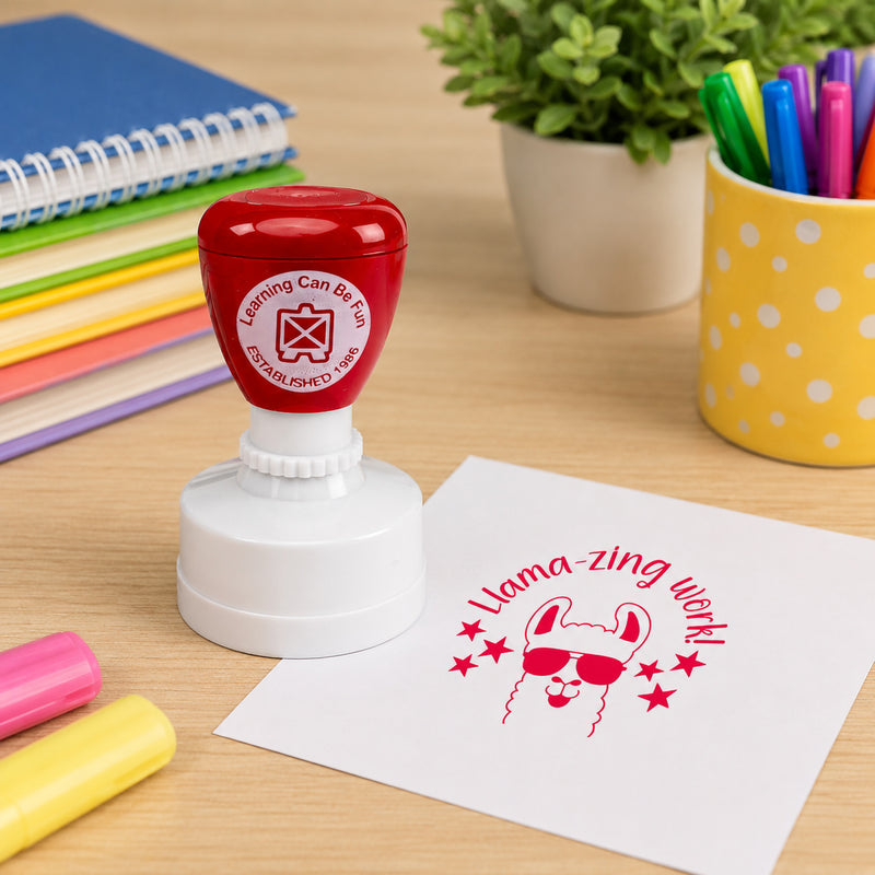 Learning Can Be Fun Llamazing Work Merit Stamp displayed on a classroom desk beside colourful markers and notebooks. The bright red llama themed impression showcases a fun motivational message that celebrates outstanding student achievement.