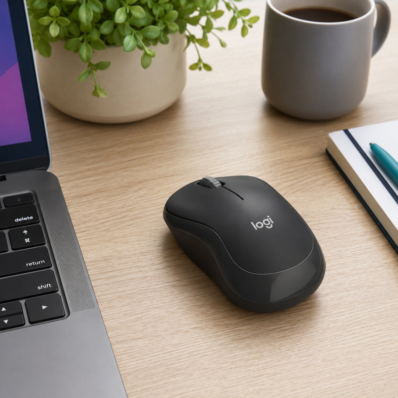 The Logitech M240 Silent Bluetooth Mouse Graphite positioned on a tidy office desk next to a laptop, notebook and coffee cup in a bright workspace, illustrating its sleek appearance and suitability for modern work environments.