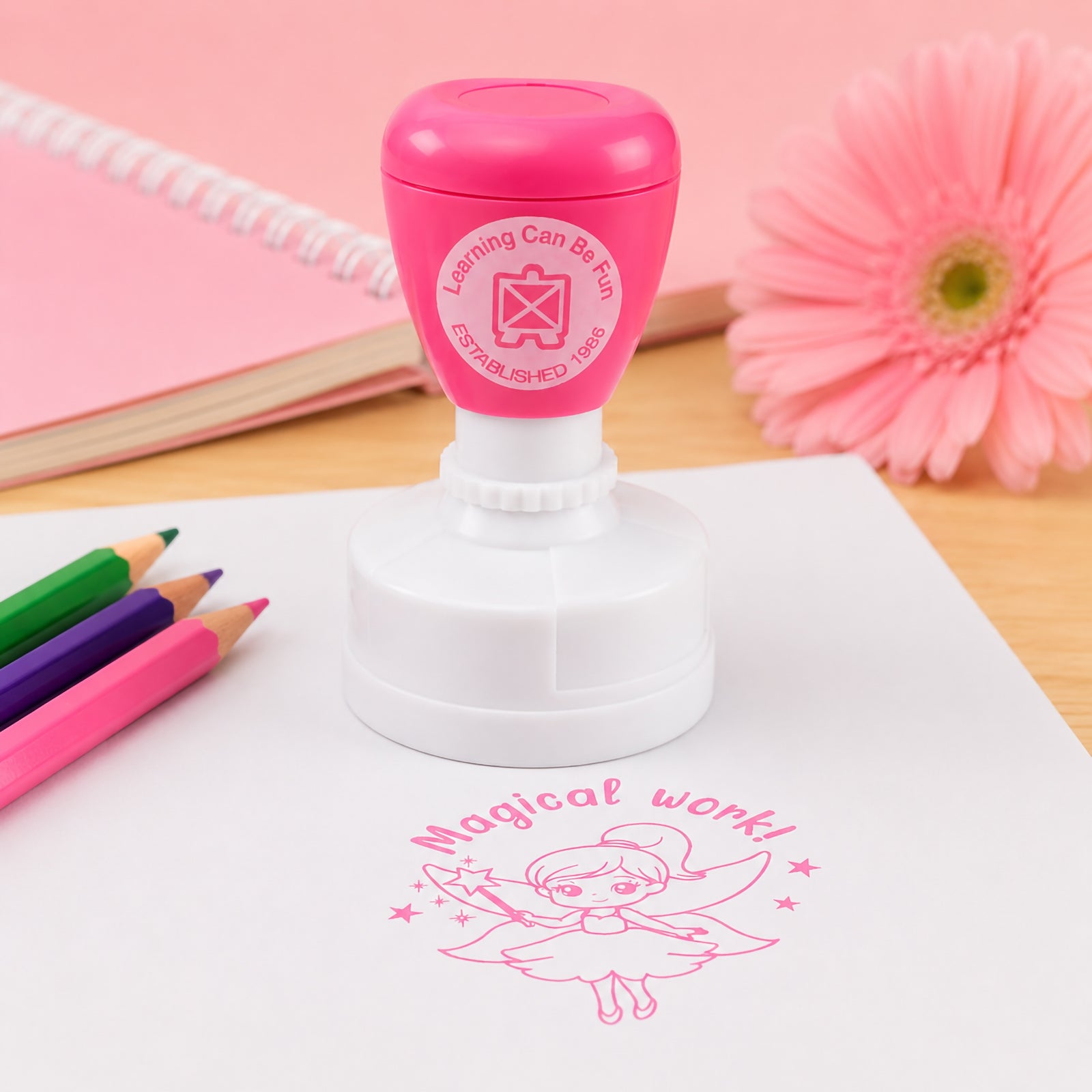 The pink self inking merit stamp is displayed on a desk surrounded by coloured pencils, stationery and a flower. A crisp fairy themed Magical Work! impression is shown beneath, illustrating the stamp's clear print quality and cheerful classroom appeal.