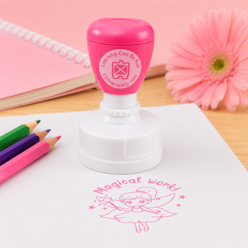 The pink self inking merit stamp is displayed on a desk surrounded by coloured pencils, stationery and a flower. A crisp fairy themed Magical Work! impression is shown beneath, illustrating the stamp's clear print quality and cheerful classroom appeal.