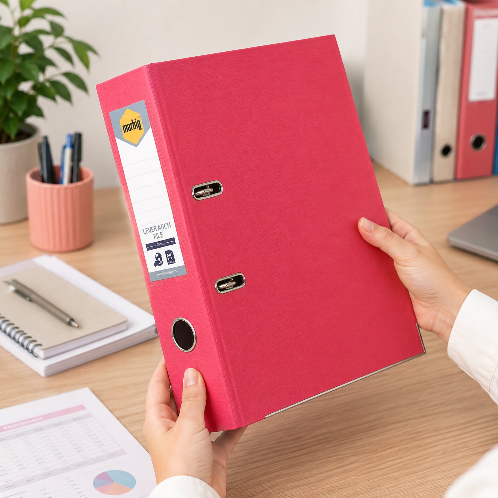 Hands holding the Marbig Lever Arch File A4 PE Coral in a contemporary office workspace, highlighting the vibrant coral finish, durable polyethylene construction and professional filing solution for everyday document organisation.