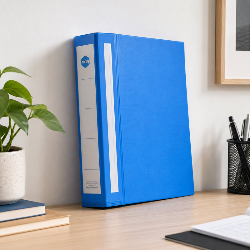 Blue Marbig Deluxe ring binder standing on a timber desk against a wall beside a plant, pens and a notebook
