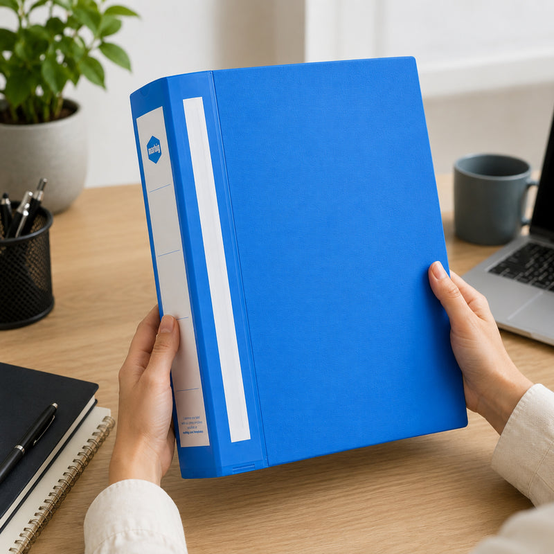 Two hands holding a blue Marbig Deluxe ring binder at a desk with a laptop and a pen cup in the background