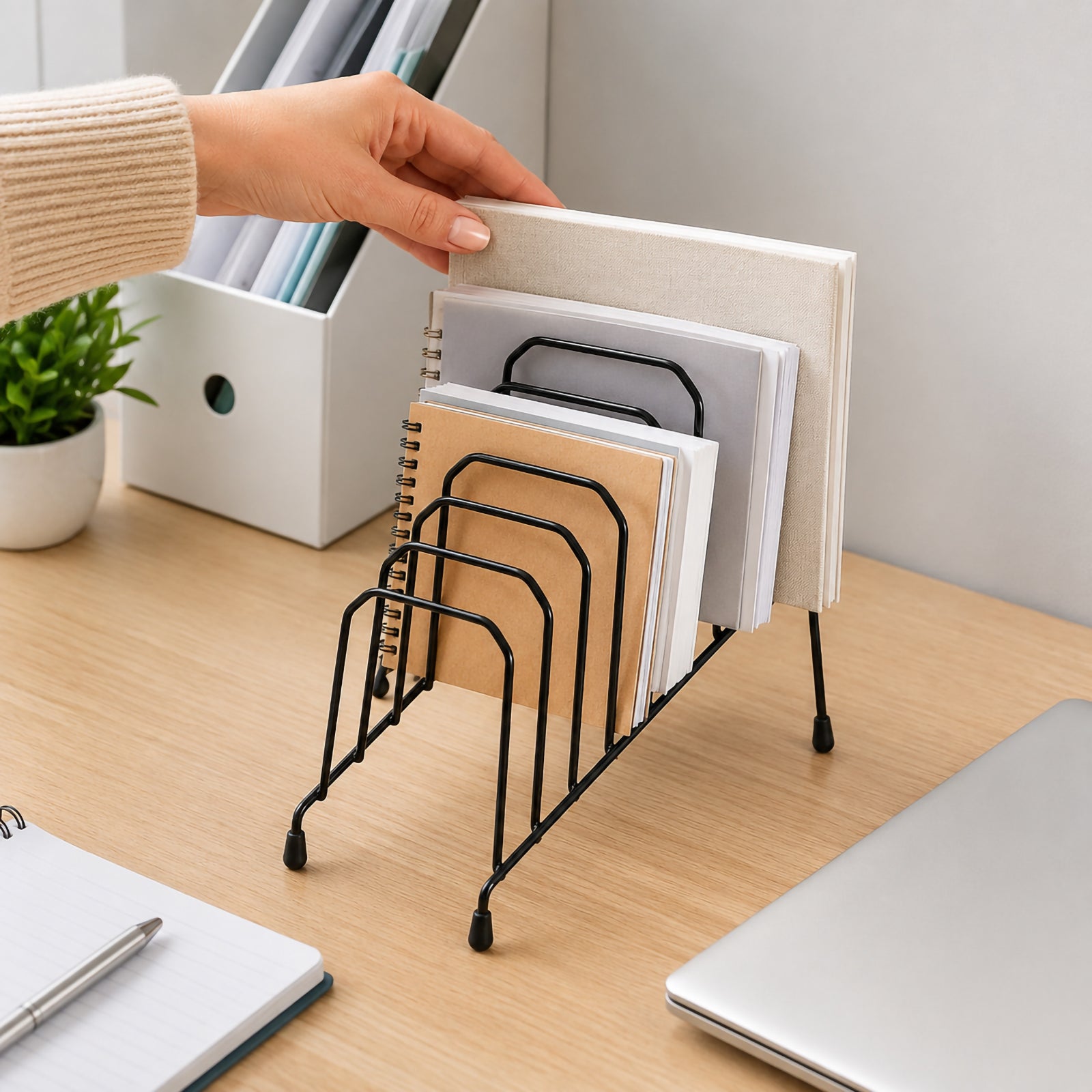 Hand placing a notebook into the Marbig Step File Wire Organiser Small Black on a modern office desk. The organiser holds multiple notebooks and folders in six graduated wire slots, demonstrating easy document access and efficient desktop organisation.