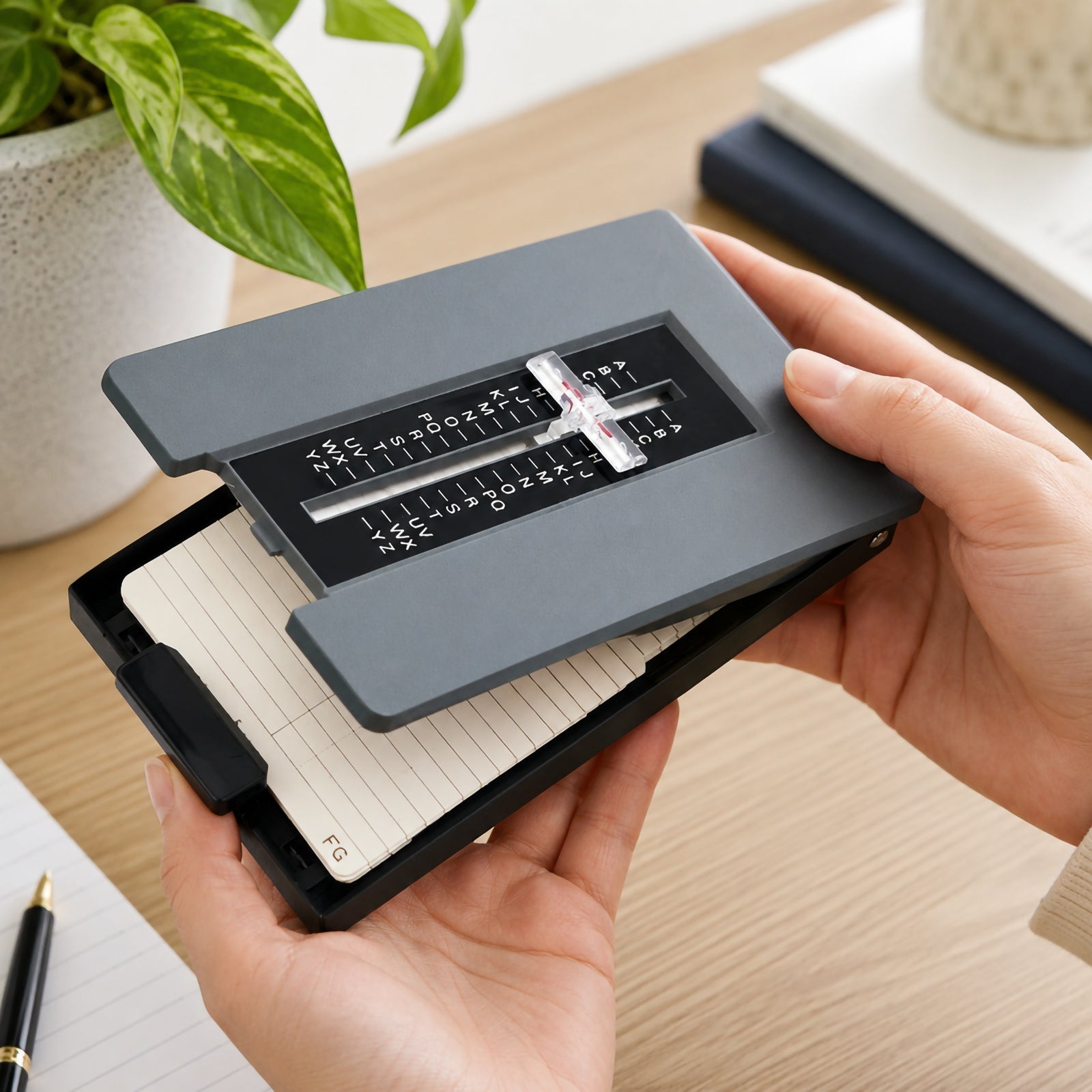 Hands holding the Marbig Telephone List Finder Grey with the cover open, showing the removable alphabetical index cards and magnifying selector in a modern office workspace with stationery and indoor plants.