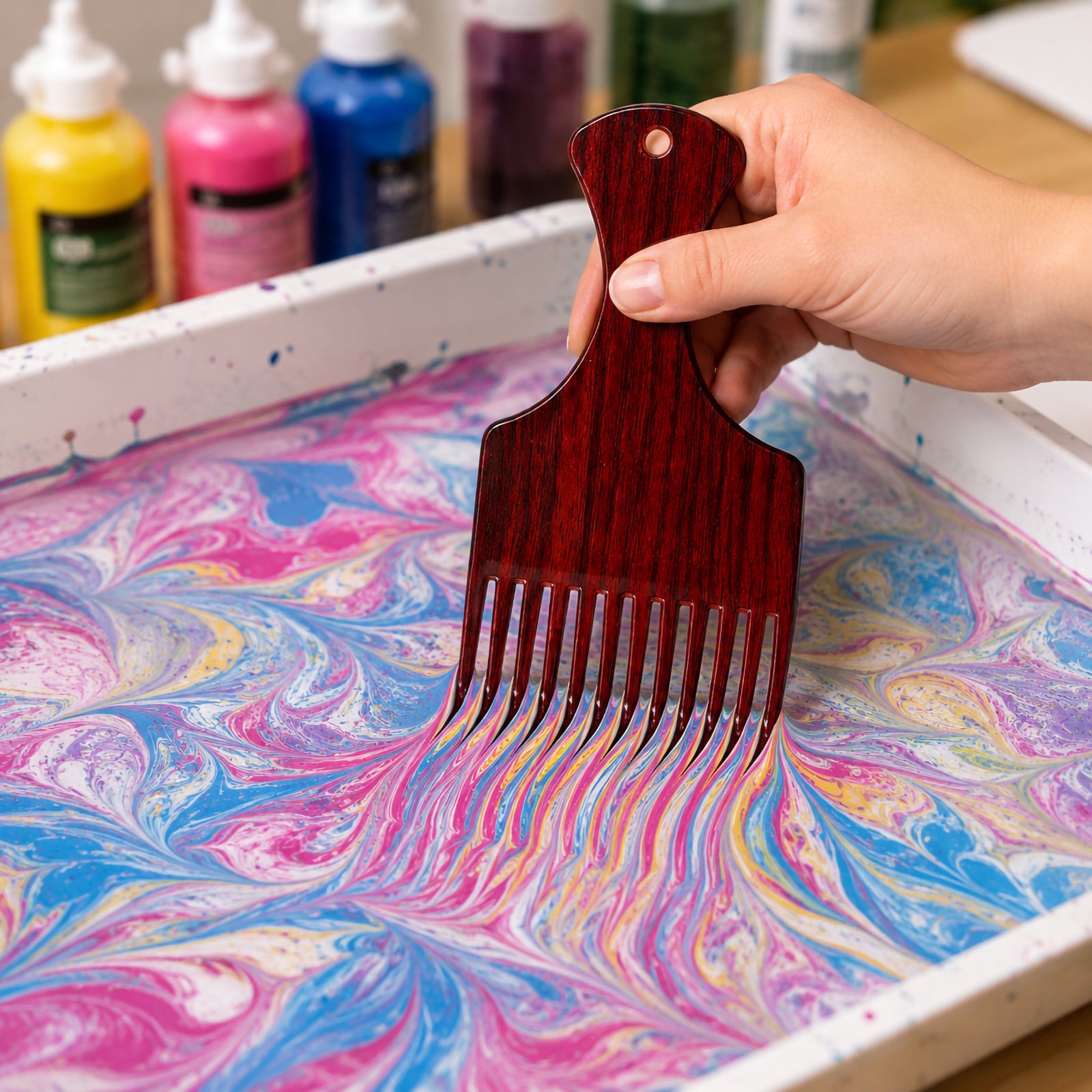 Close up of a hand using the marbling ink comb to create flowing pink, blue, and yellow marbled paint patterns in a shallow art tray.