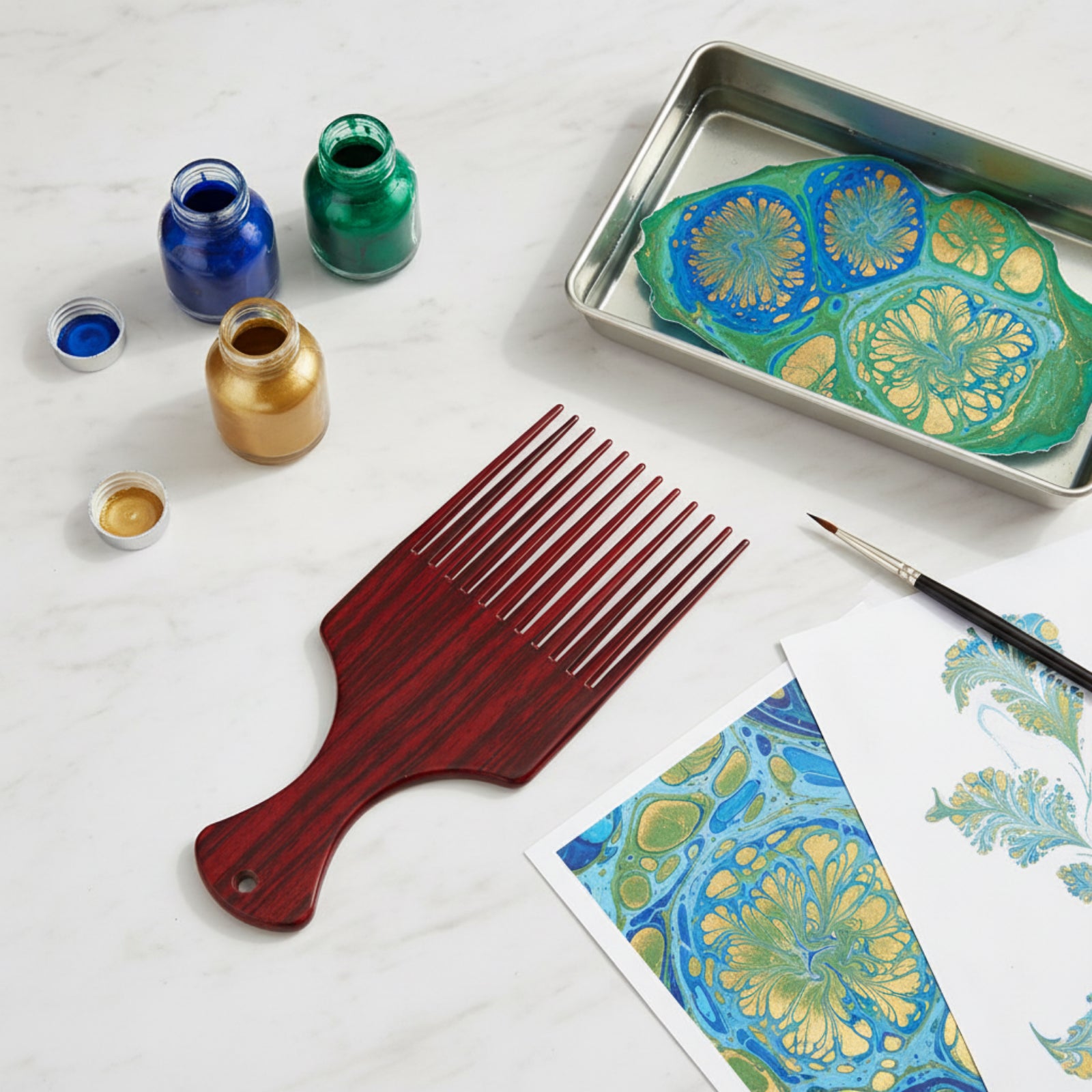 Marbling Ink Comb positioned next to metallic blue and gold marbling inks with detailed marbled artwork and creative paper designs for art projects.