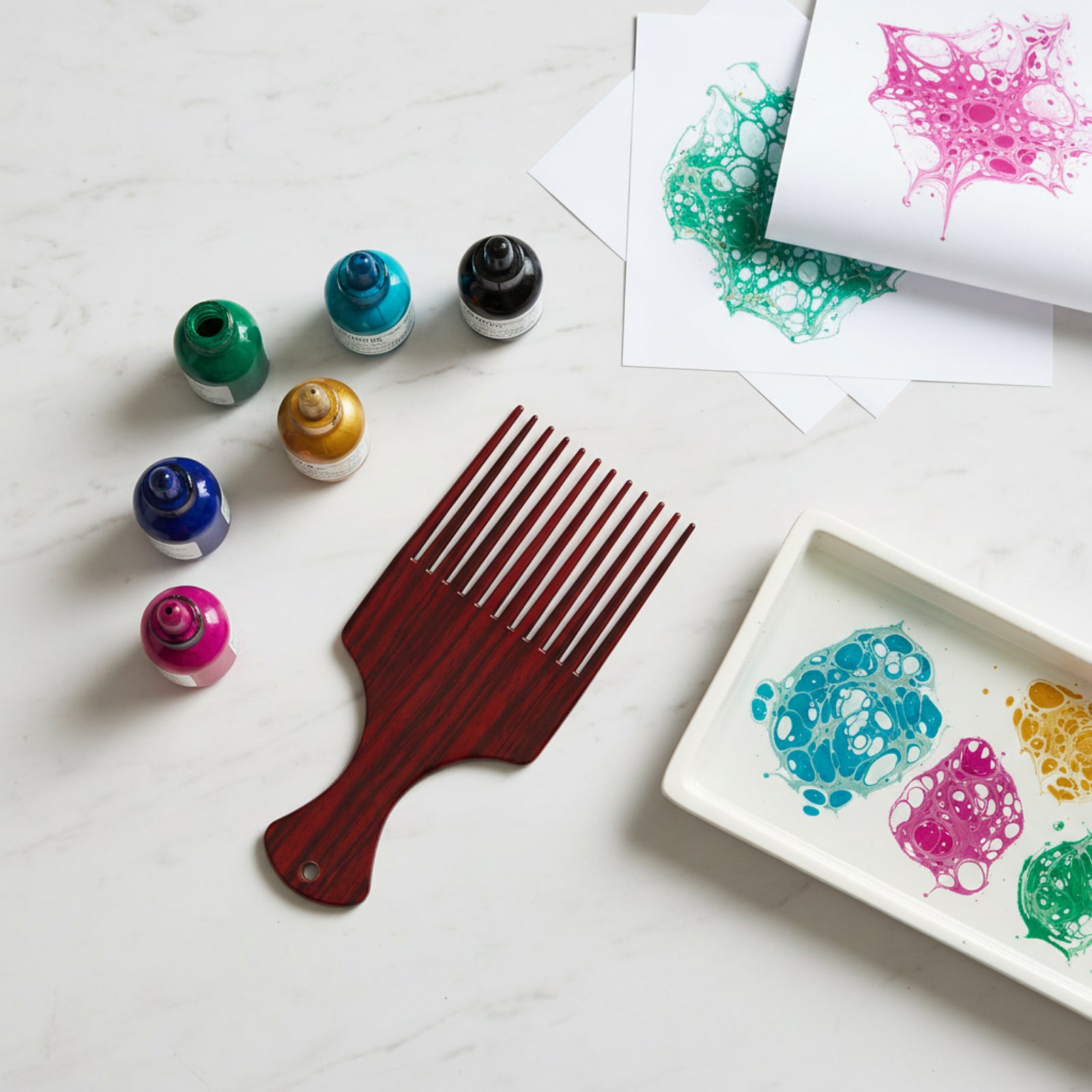 Educational Colours Marbling Ink Comb displayed beside colourful marbling inks, marbled paper samples, and a tray of decorative paint patterns on a white tabletop.