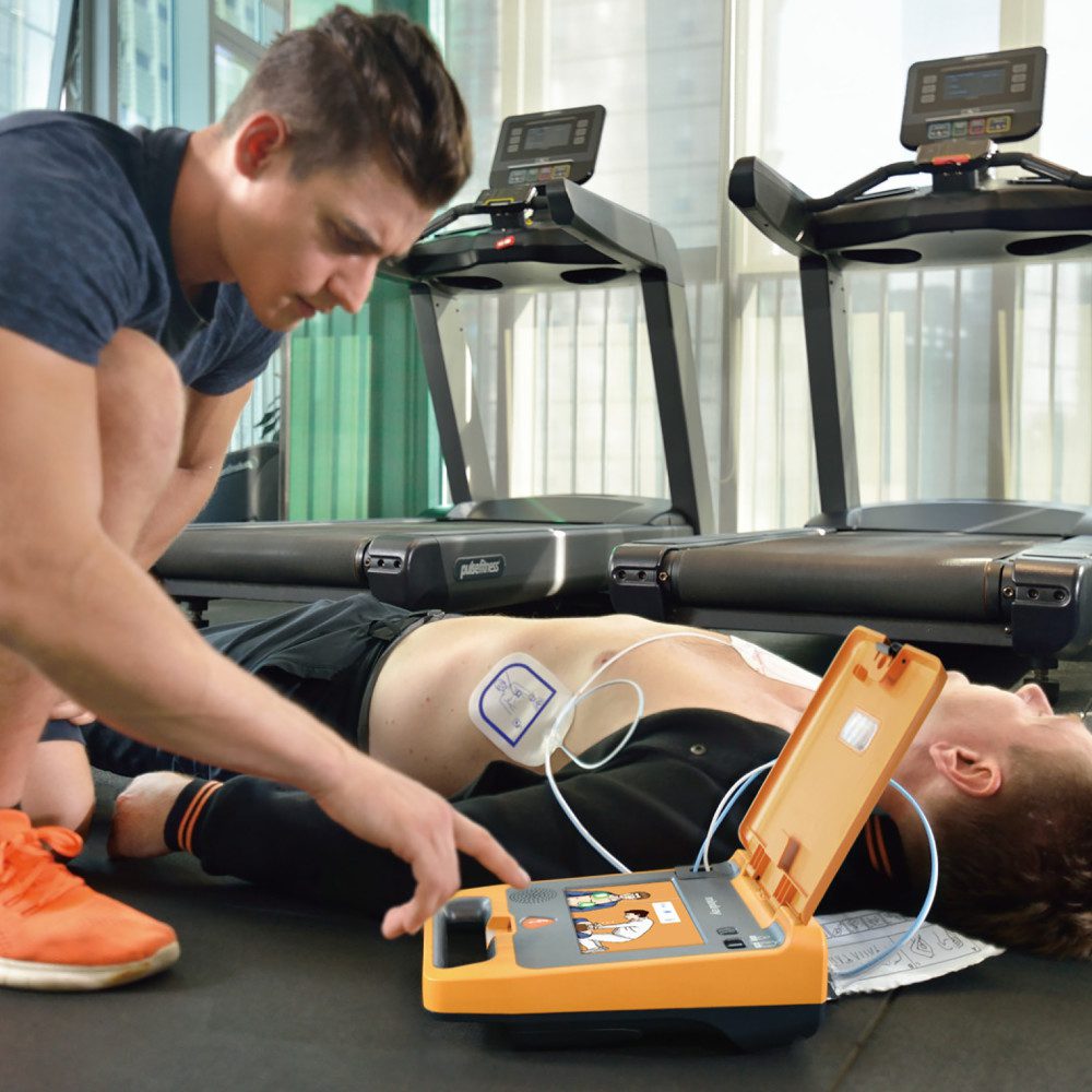 Rescuer kneeling beside a patient in a gym with a Mindray C1A open and pads applied to the chest