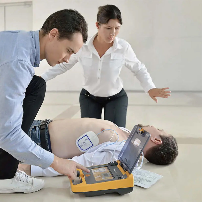 Mindray C2 Fully Automatic Defibrillator in use during a simulated emergency with electrode pads attached to an adult patient while two responders follow the device guidance and coordinate the cardiac arrest response.