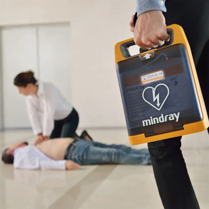 Mindray C2 Fully Automatic Defibrillator being carried rapidly towards an indoor casualty while another person begins emergency assistance. The integrated carry handle and compact case demonstrate how the AED can be moved quickly from its storage point to the emergency scene.