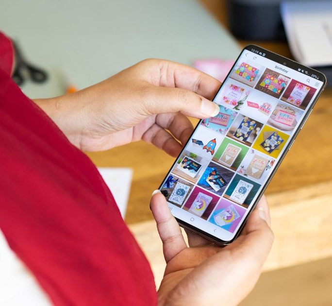A person holding a black smartphone with both hands, showing a photo gallery screen on the device. The person is wearing a red top, and the background includes a wooden table and a blurred indoor setting.