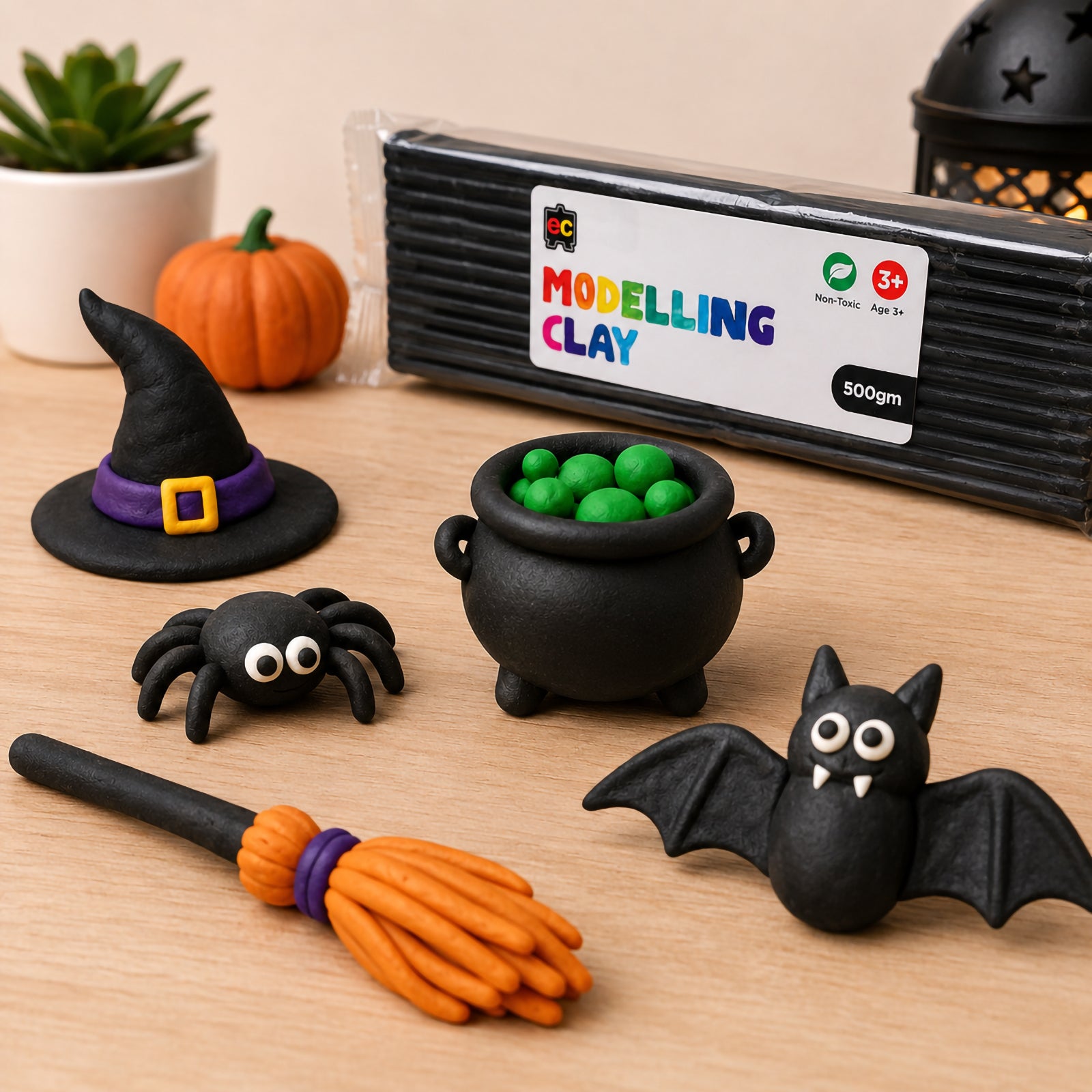 Educational Colours 500g black modelling clay displayed on a wooden craft table with handmade Halloween clay figures including a bat, spider, witch hat, cauldron and broomstick in a warm lifestyle setting.
