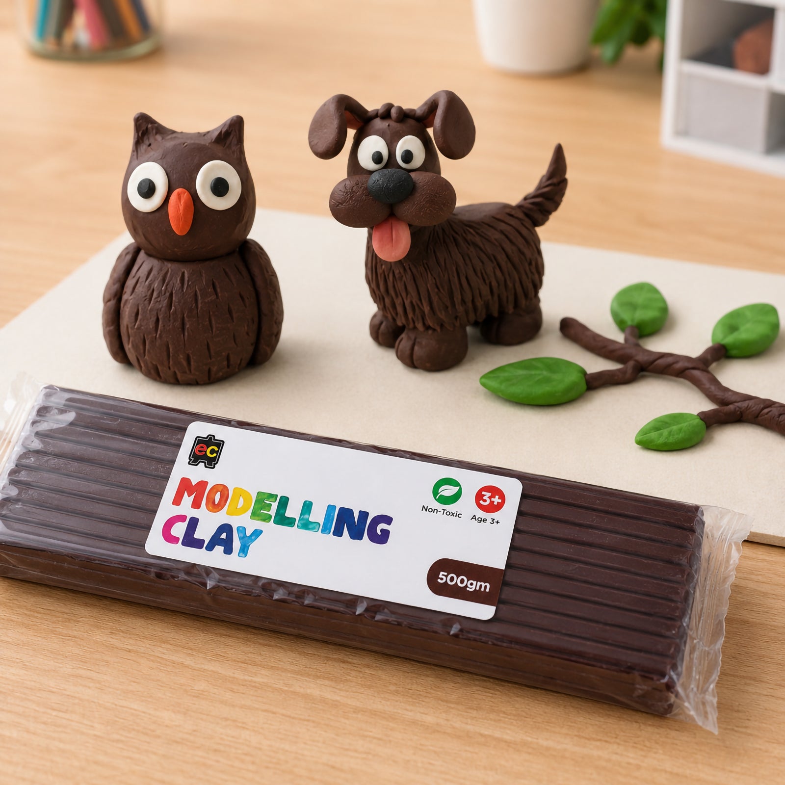 Brown modelling clay pack with handcrafted clay owl, dog, and green leaf branch arranged on a clean tabletop in a softly lit arts and crafts setting.
