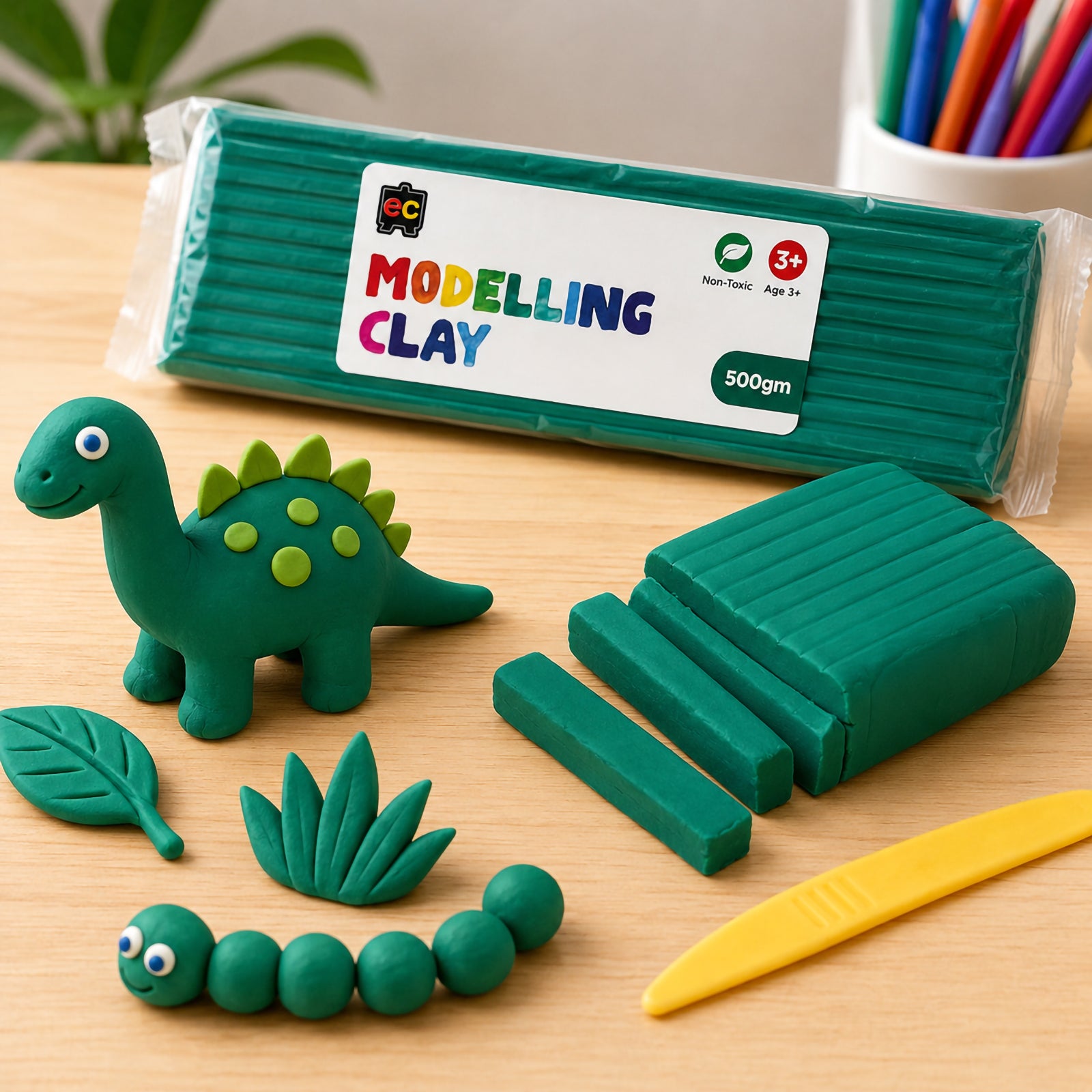 Educational Colours 500g dark green modelling clay displayed on a wooden craft table with a handmade green clay dinosaur, clay leaves, sculpting tool, and sliced clay pieces in a bright classroom-style setting.