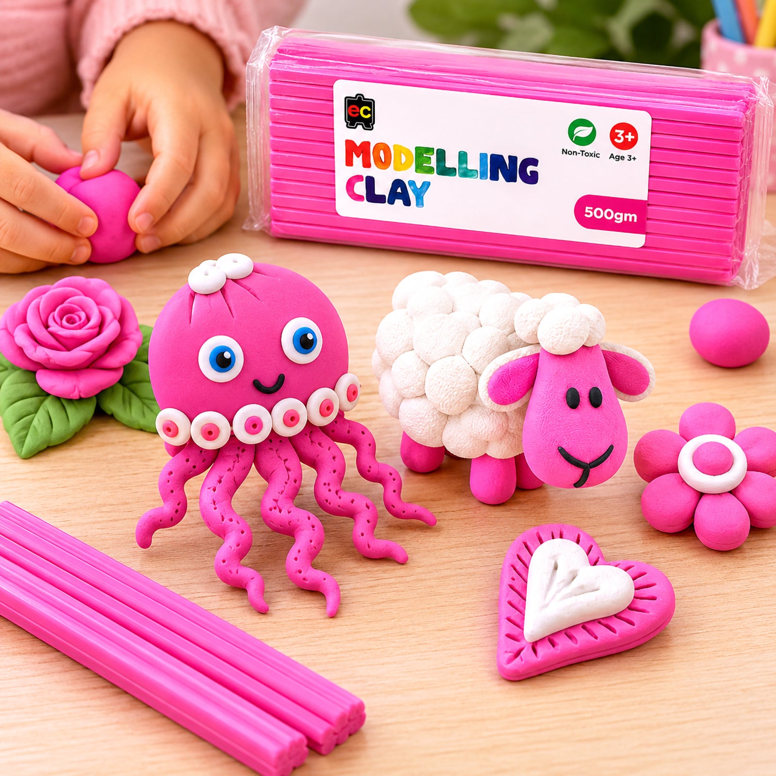 Child shaping pink modelling clay beside handmade clay octopus, sheep, flower, and heart creations with Educational Colours Modelling Clay 500g Pink packaging on a wooden table.