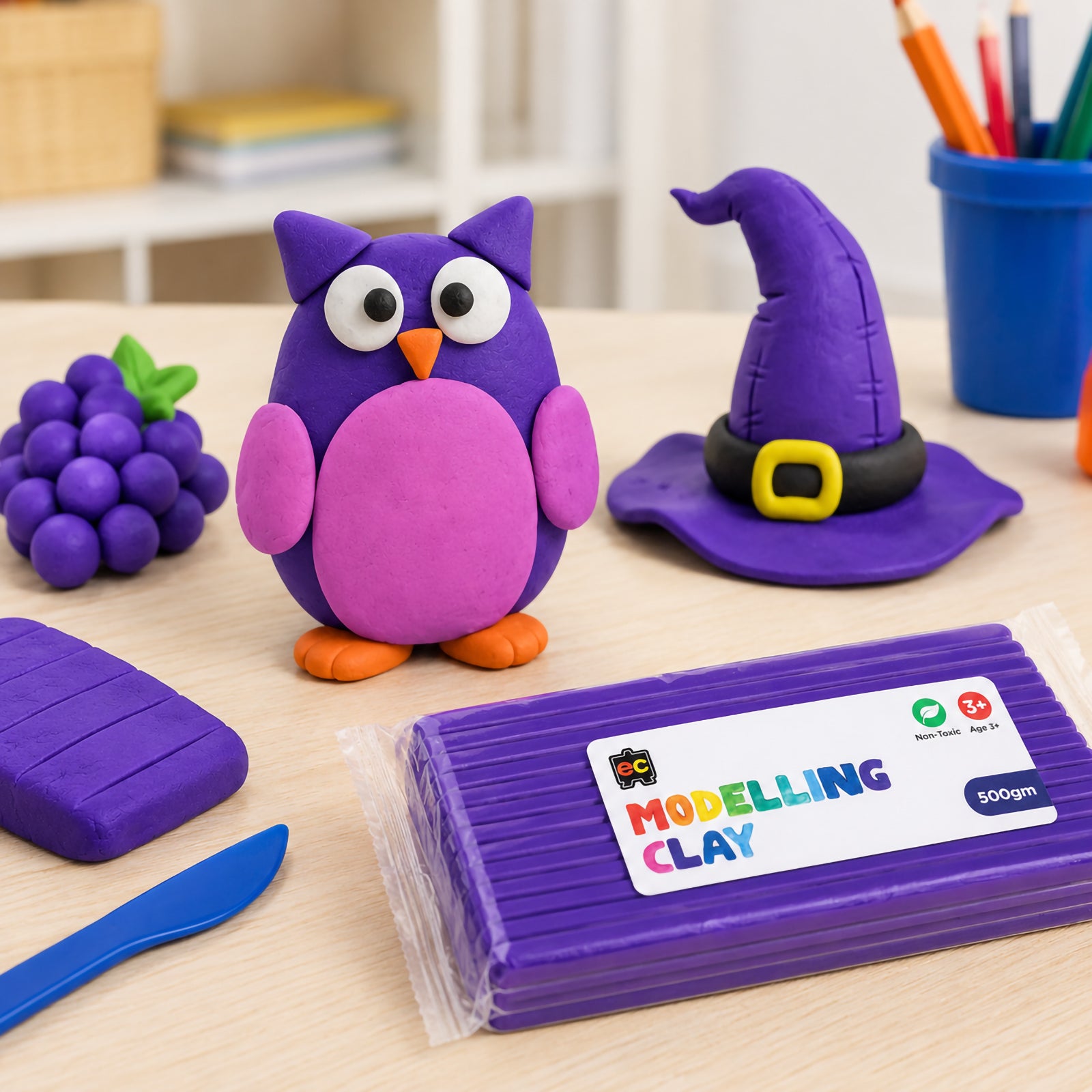 Purple modelling clay owl, witch hat, and grape sculptures displayed on a light wooden craft table beside Educational Colours Modelling Clay 500g Purple packaging and sculpting tools.