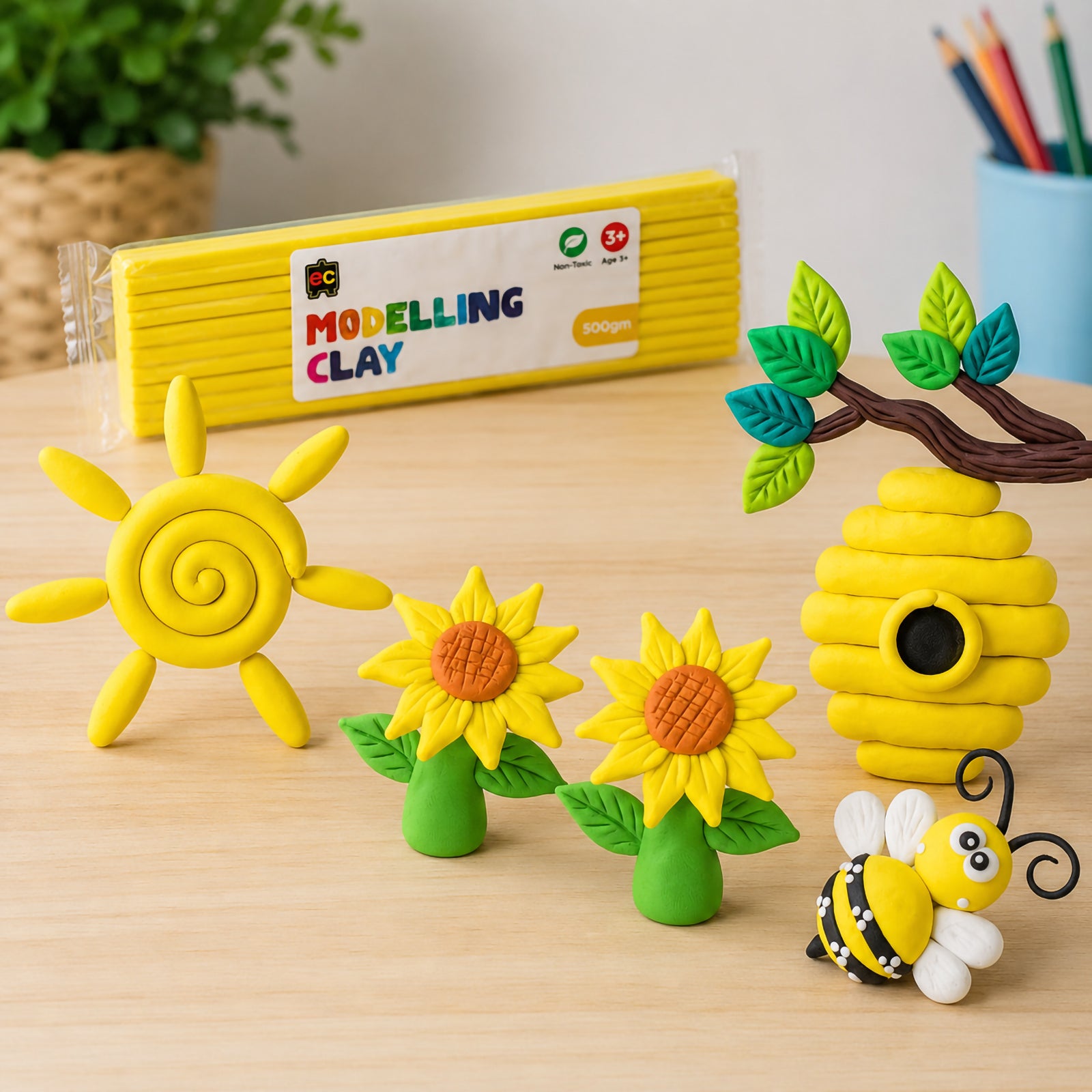 Yellow modelling clay craft display featuring a spiral sun, sunflower flowers, bee hive and bee arranged on a wooden tabletop beside Educational Colours 500g yellow modelling clay packaging.