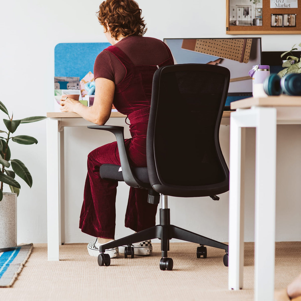 Person seated on a black Mondo Fleet mesh chair at a white desk in an office, rear view showing the mesh back and fixed armrests