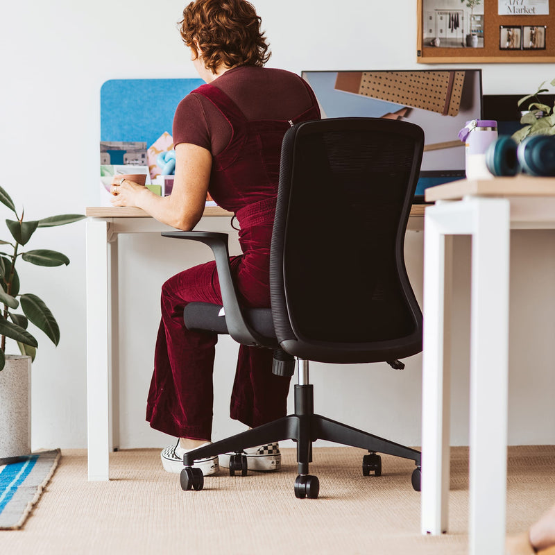 Person seated on a black Mondo Fleet mesh chair at a white desk in an office, rear view showing the mesh back and fixed armrests