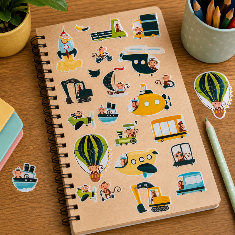 Kraft notebook decorated with Monkey Adventures foil stickers featuring cheerful monkeys exploring the world through different vehicles and modes of transport. Hot air balloons, submarines, aeroplanes and construction vehicles create a playful adventure scene on a wooden desk.