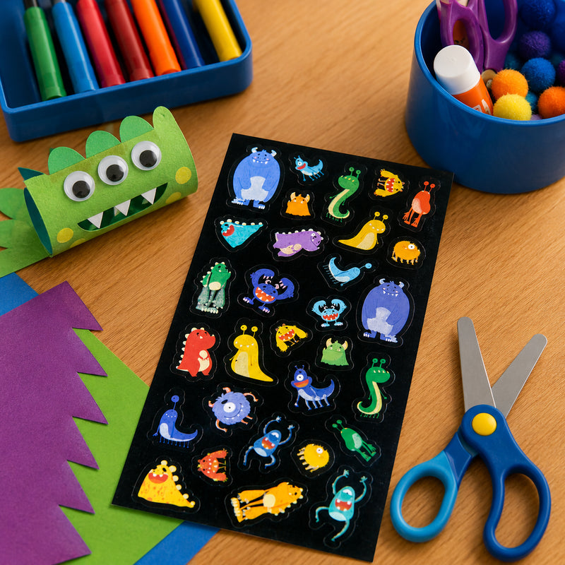 Complete sticker sheet displayed on a craft table with colourful paper, scissors and handmade monster crafts. The collection features cheerful monsters in a variety of shapes, colours and playful expressions.