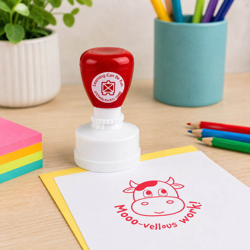 Classroom workspace featuring the red merit stamp with a clear Mooo vellous Work impression on paper. Colourful pencils and sticky notes highlight its use as a motivational teaching tool.