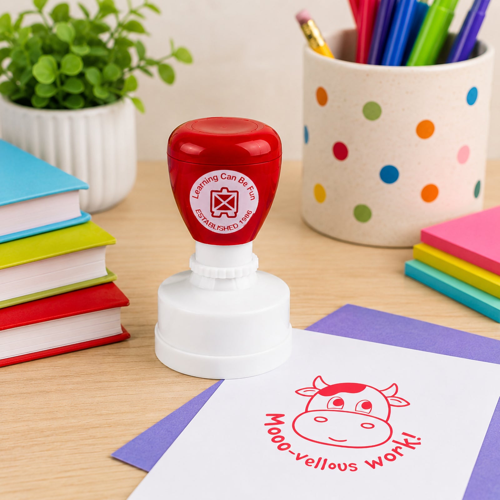 Learning Can Be Fun Mooo vellous Work Merit Stamp displayed on a classroom desk surrounded by colourful books, stationery and learning resources. The bright red cow themed stamp impression provides a fun way to recognise excellent student work.