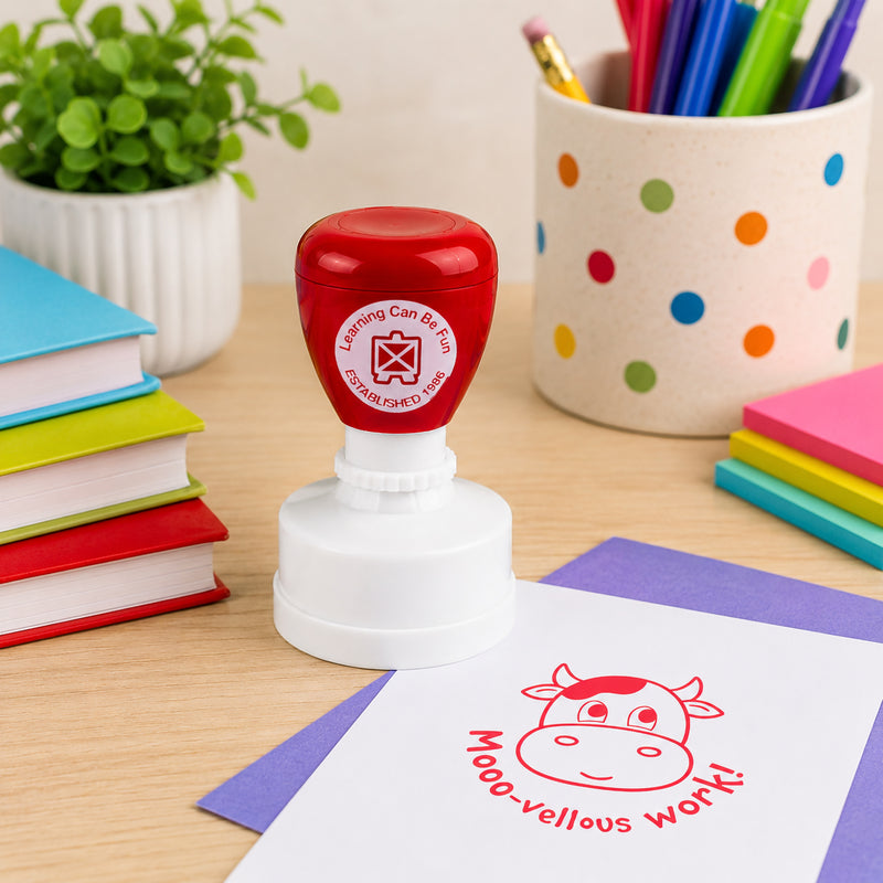 Learning Can Be Fun Mooo vellous Work Merit Stamp displayed on a classroom desk surrounded by colourful books, stationery and learning resources. The bright red cow themed stamp impression provides a fun way to recognise excellent student work.