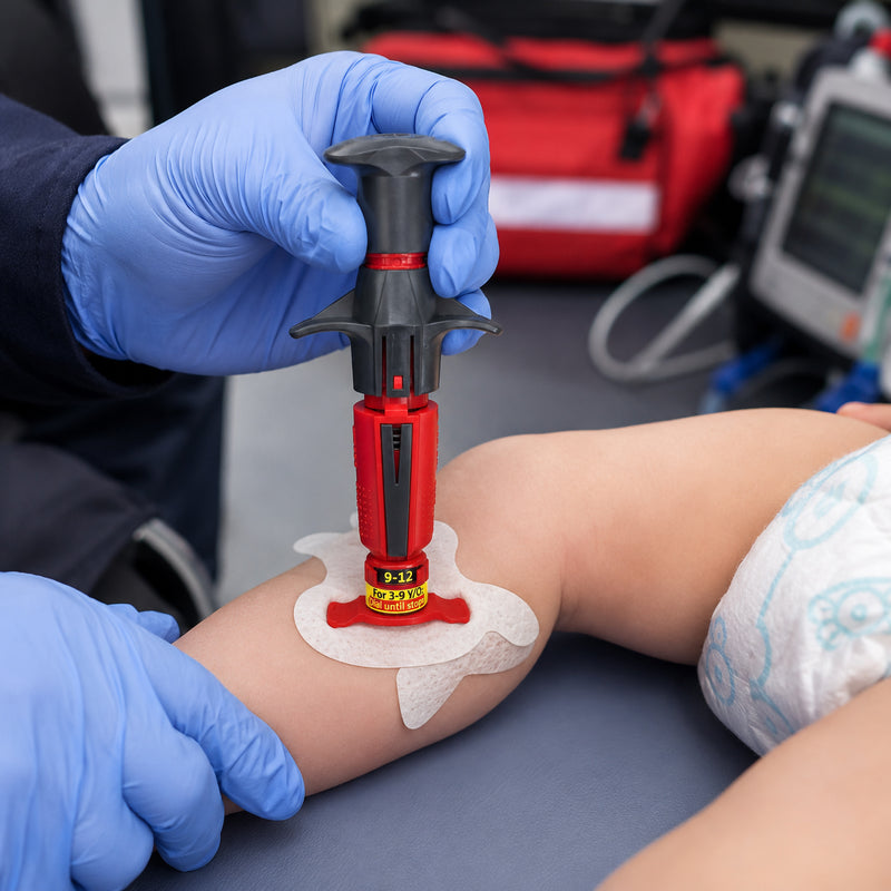 NIO Infant Intraosseous Device positioned on an infant training mannequin leg during a simulated emergency procedure. Blue gloved clinical hands support the limb while the red and black automatic IO device is shown with a fixation dressing around the placement area. Emergency medical equipment appears softly in the background, illustrating a controlled clinical training environment for infant vascular access.