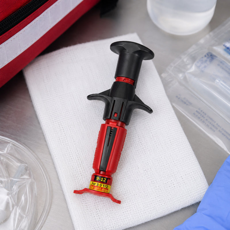NIO Infant Intraosseous Device displayed from above on a folded sterile dressing beside emergency medical supplies. The red and black spring loaded device is shown clearly against the clean clinical surface, with a medical response bag, packaged accessories and preparation materials surrounding it to demonstrate readiness for emergency care equipment organisation.