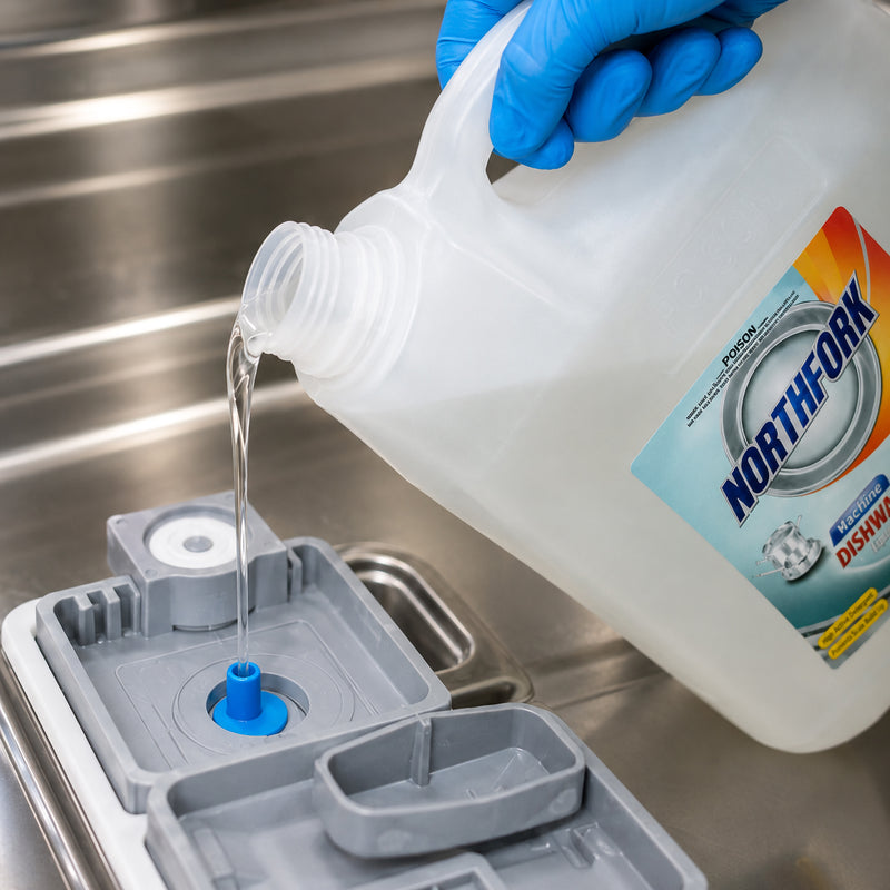 Northfork Machine Dishwashing Liquid 5L being carefully poured into the detergent reservoir of a commercial dishwasher by a gloved operator. The image highlights the correct filling method using an automatic dispensing system within a professional stainless steel kitchen environment.