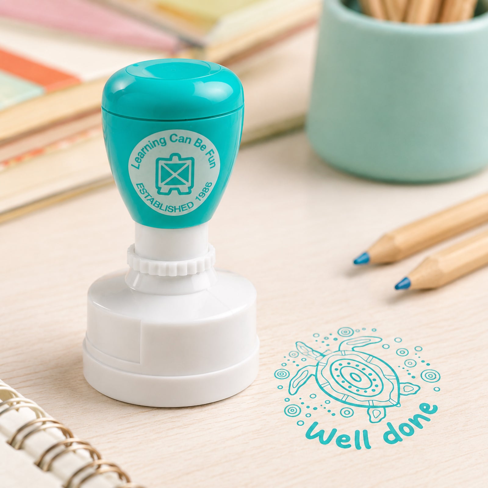 The turquoise self inking merit stamp stands beside notebooks and coloured pencils, displaying a crisp turtle themed Well Done impression. The classroom styled setting showcases the stamp as a practical and engaging tool for everyday educational use.