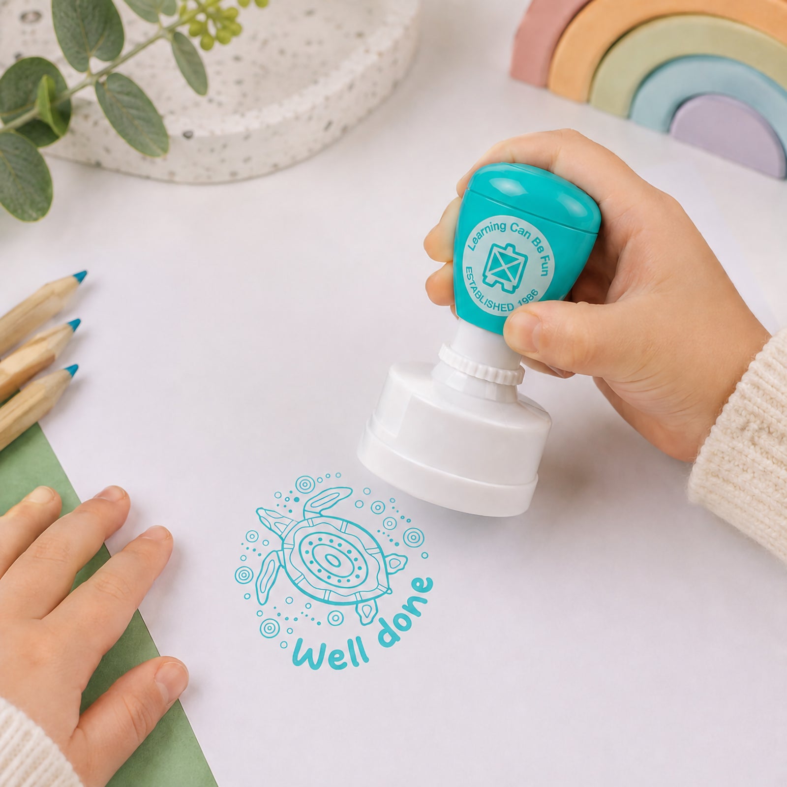 A child uses the Ocean Country Turtle Merit Stamp on a sheet of paper, creating a clear turquoise turtle impression with the Well Done message. The image highlights the easy self inking design and demonstrates how teachers can quickly reward student effort with positive feedback.