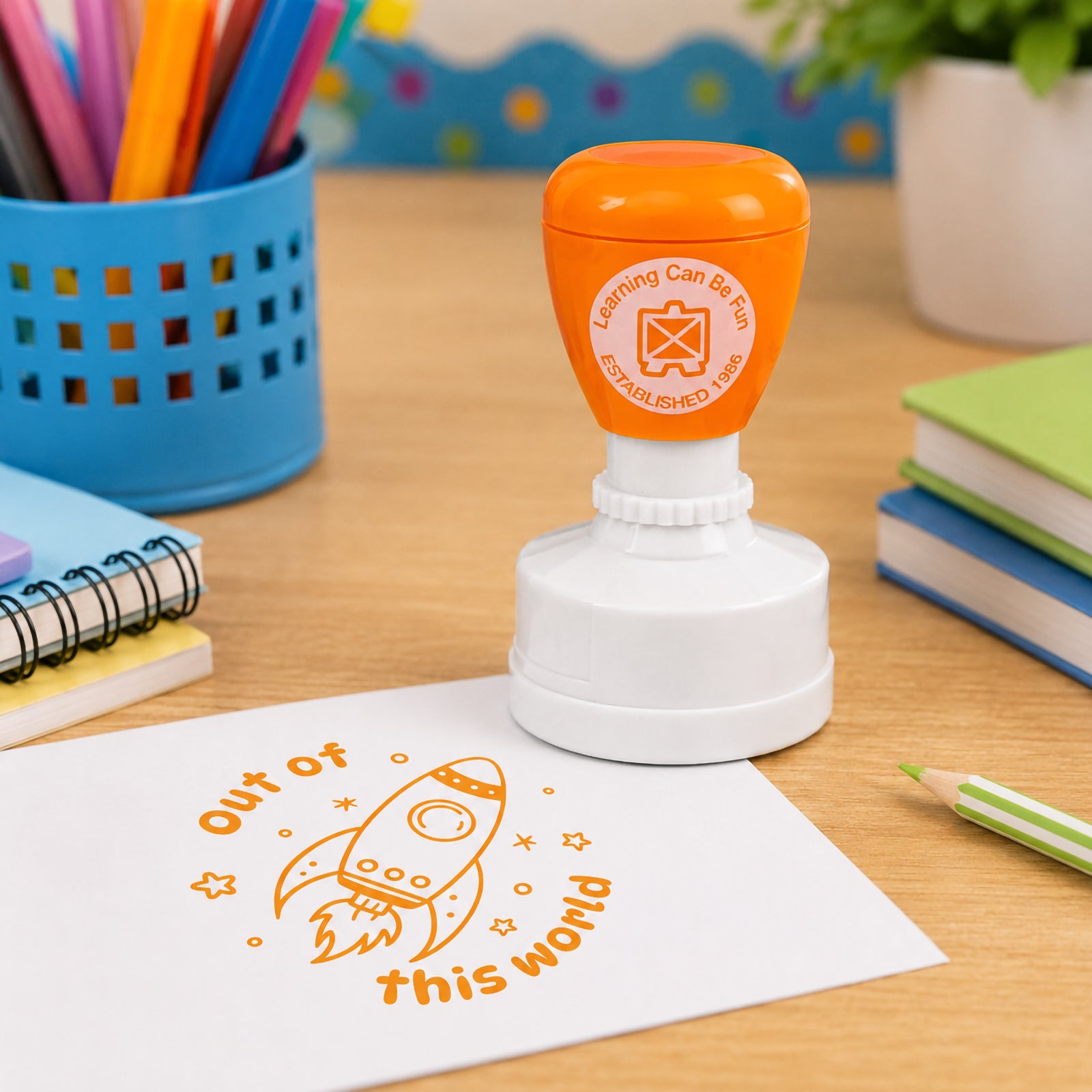 Self inking merit stamp positioned on a classroom desk with coloured pencils and exercise books nearby. The vibrant orange Out of This World impression highlights positive feedback and encourages students to reach their learning goals.
