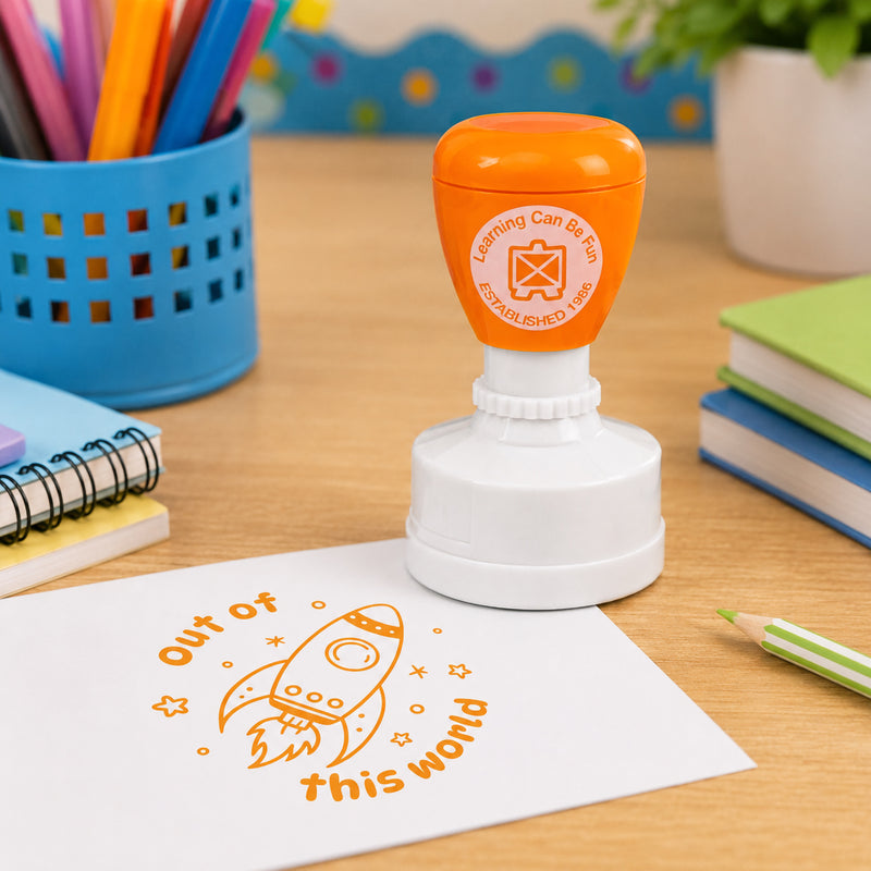 Self inking merit stamp positioned on a classroom desk with coloured pencils and exercise books nearby. The vibrant orange Out of This World impression highlights positive feedback and encourages students to reach their learning goals.