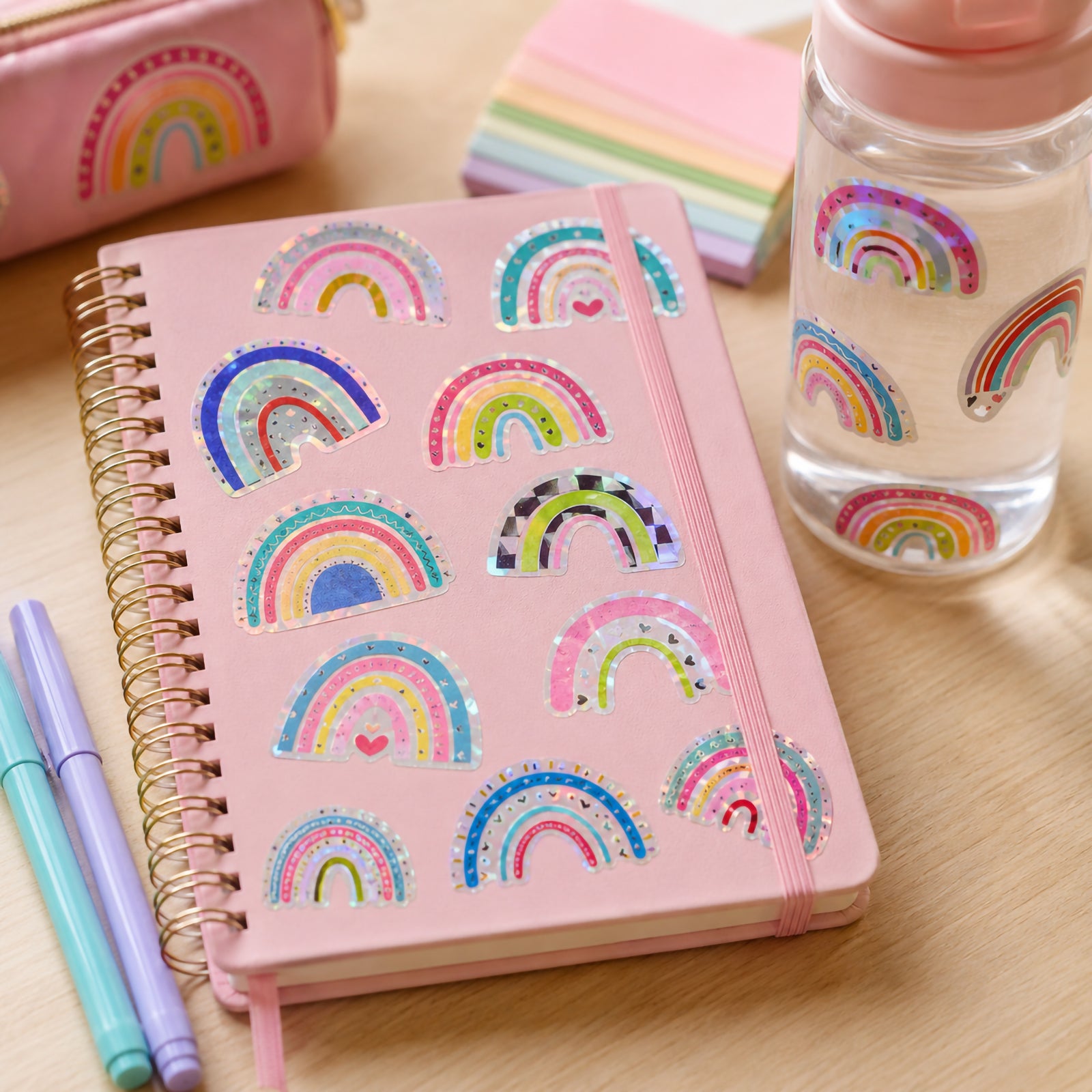 Close up of a pink spiral notebook and water bottle decorated with holographic rainbow stickers. Various rainbow patterns shimmer under natural light, highlighting the colourful and playful designs.