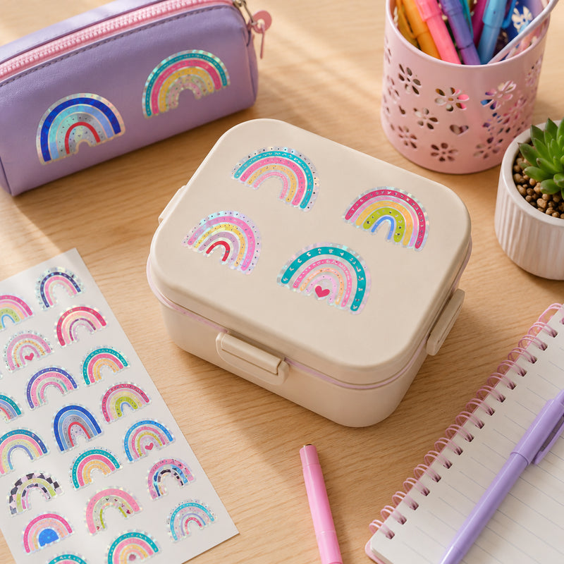 Over the Rainbow holographic stickers decorating a lunch box, pencil case and stationery items. Multiple rainbow designs showcase vibrant colours and reflective foil finishes that add brightness to everyday accessories.