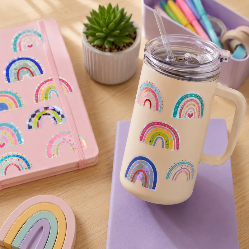 Pastel rainbow holographic stickers displayed on a reusable travel mug and pink notebook. The colourful rainbow designs feature sparkling foil details and decorative patterns, creating a cheerful and uplifting stationery theme.