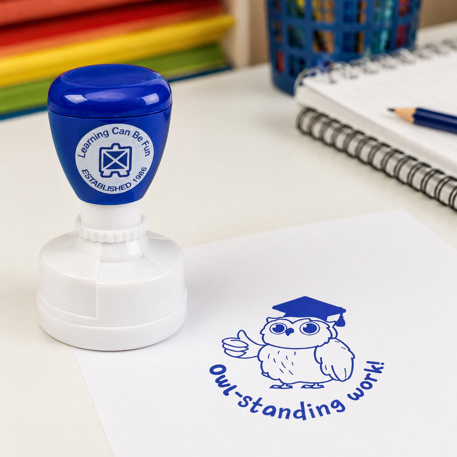 Self inking teacher reward stamp positioned next to worksheets and classroom supplies. The bold blue Owl-standing Work design creates clear and encouraging feedback that celebrates academic excellence and motivates continued learning success.