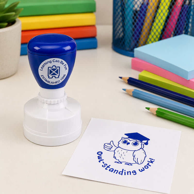 Learning Can Be Fun Owl-standing Work Merit Stamp displayed on a classroom desk beside coloured pencils, notebooks and stationery. The blue owl themed impression features a graduation cap and positive reward message, making it ideal for recognising exceptional student achievement.