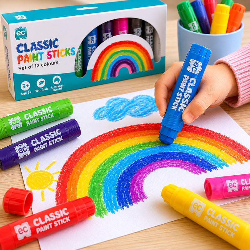 Close-up of a child colouring a rainbow in a sketchbook using Educational Colours Classic Paint Sticks, with assorted paint sticks displayed in a desktop holder.