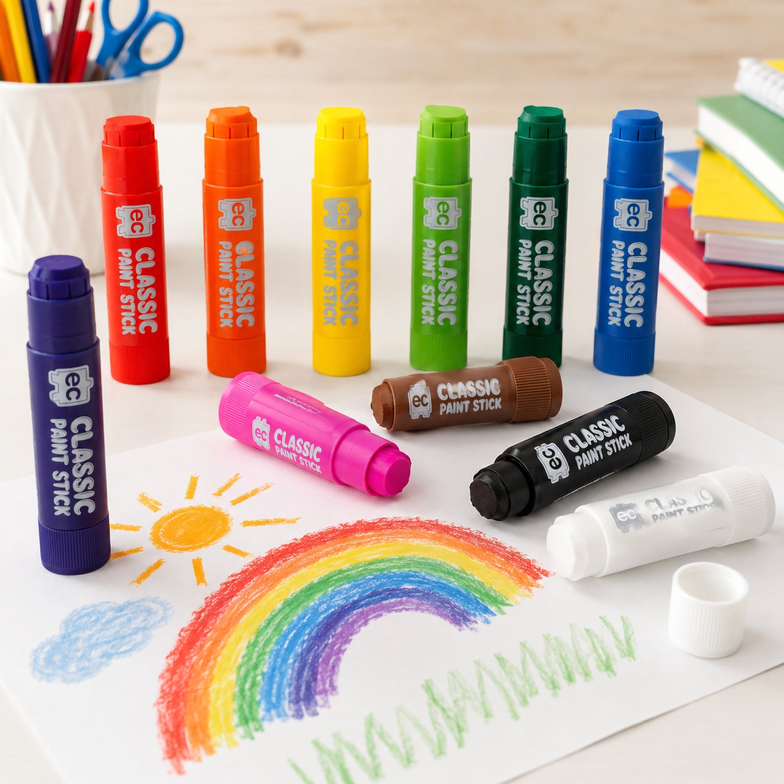 Child using a blue Educational Colours Classic Paint Stick to draw a rainbow on paper, surrounded by colourful paint sticks and the Classic Paint Sticks Set of 12 box on a wooden table.