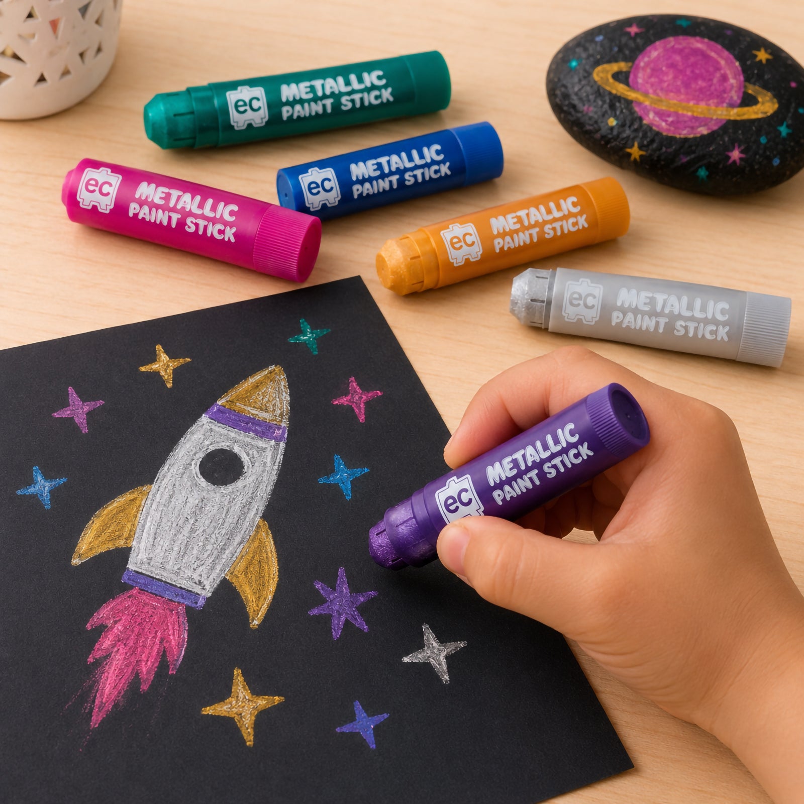 Metallic paint sticks arranged on a wooden table beside black paper featuring a colourful rocket drawing and space-themed painted rock activity.