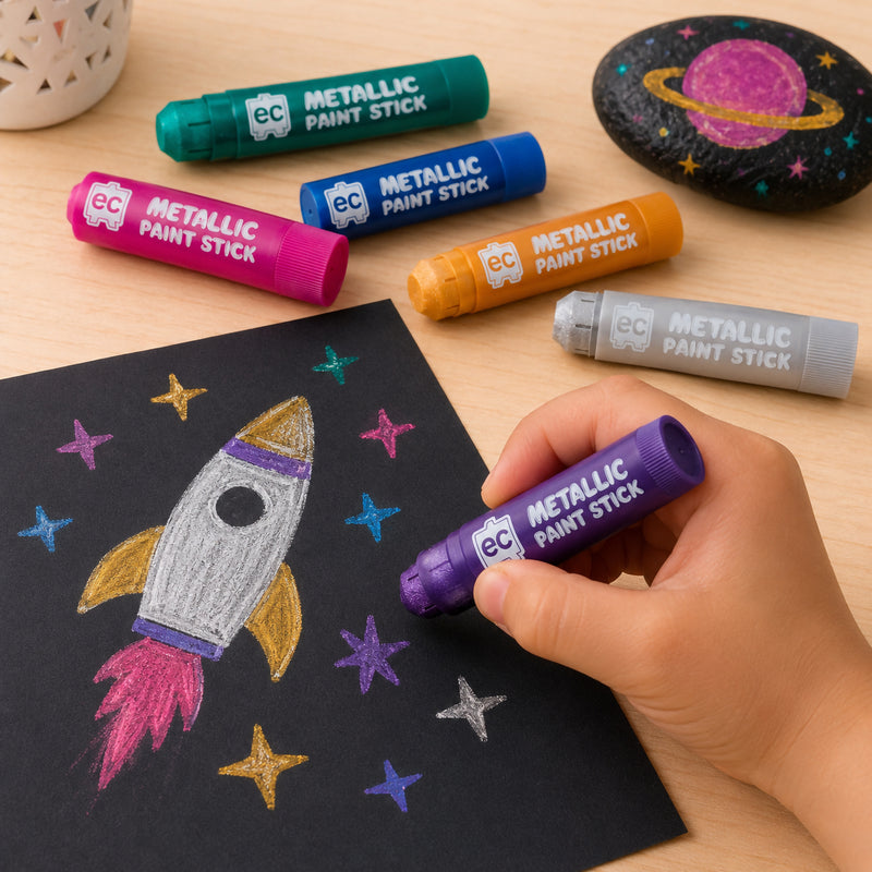 Metallic paint sticks arranged on a wooden table beside black paper featuring a colourful rocket drawing and space-themed painted rock activity.